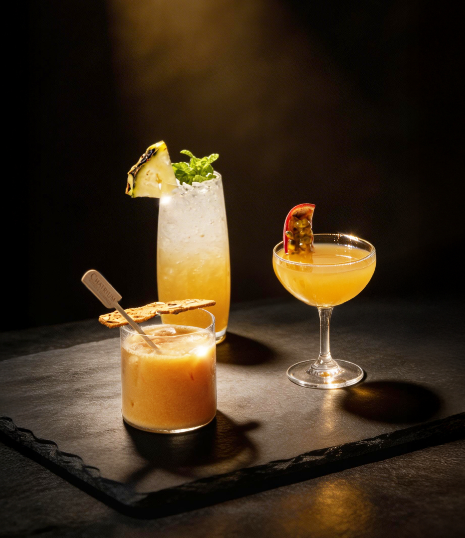 Three tropical cocktails on a slate board: pineapple-mint highball with crushed ice, creamy orange rocks with a biscuit, and a passion-fruit coupe.