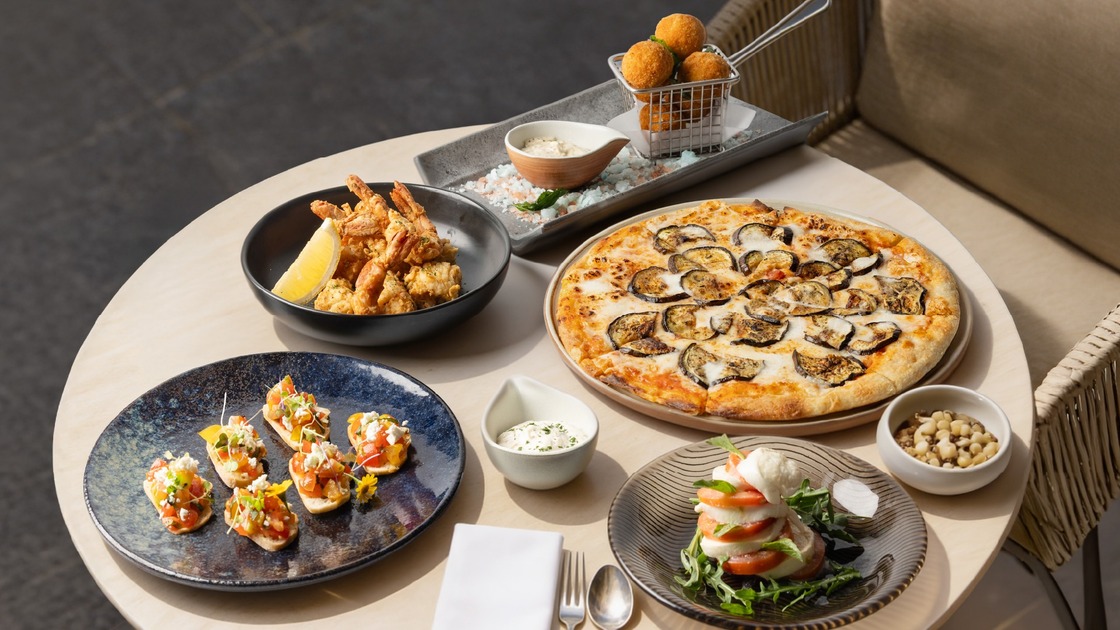 Alfresco shared dining: eggplant pizza, bruschetta, fried prawns with lemon, caprese stack and croquette bites served on a round hotel terrace table.