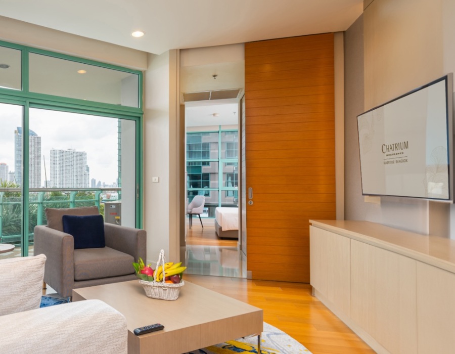 Contemporary suite living area: sofa, armchair, coffee table with fruit basket, balcony view, sliding wood door to bedroom