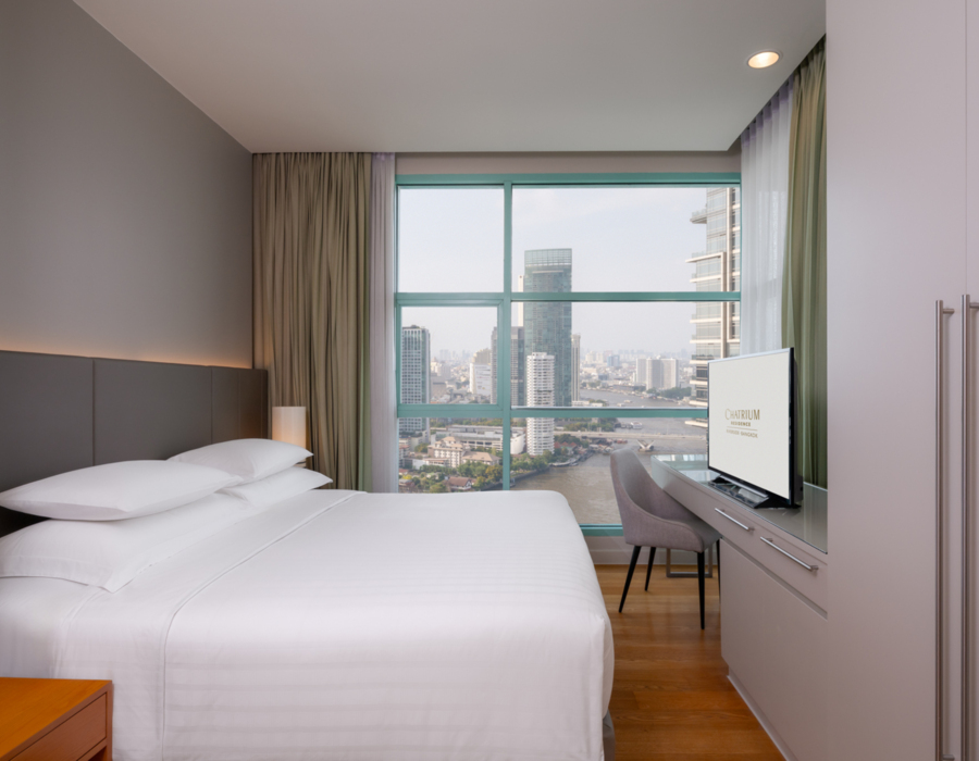 A well-furnished bedroom with a bed, a television on a stand, and a window providing a river view