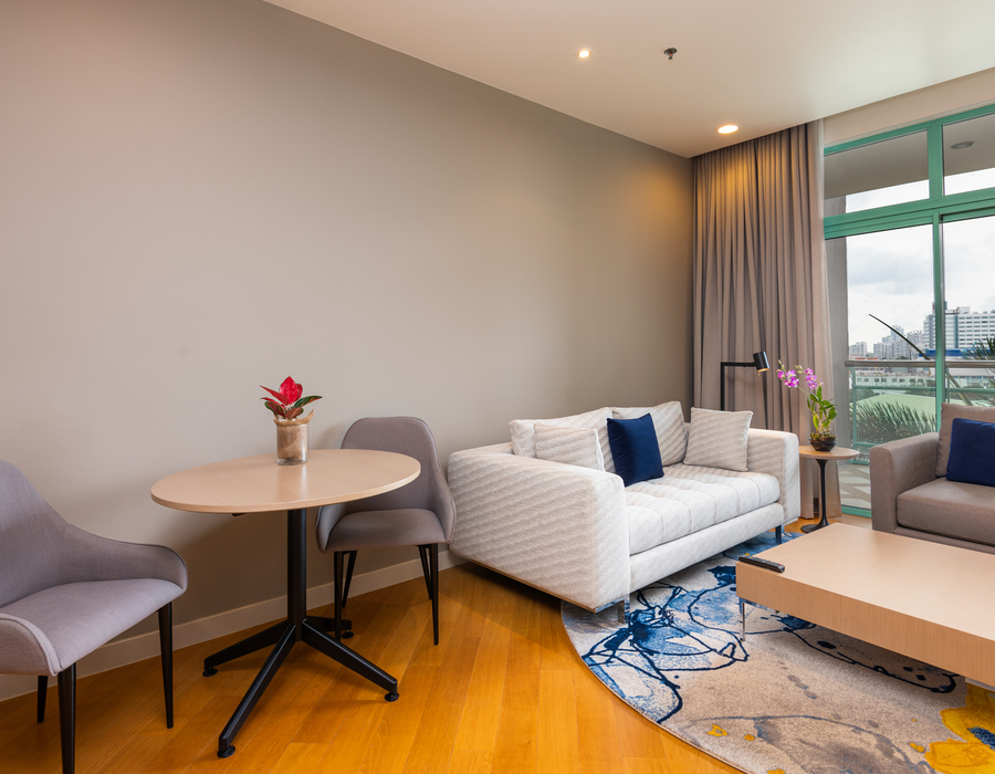 Modern hotel suite living area with sofa, armchair, round dining table, balcony city view, decorative rug and blue accent cushions