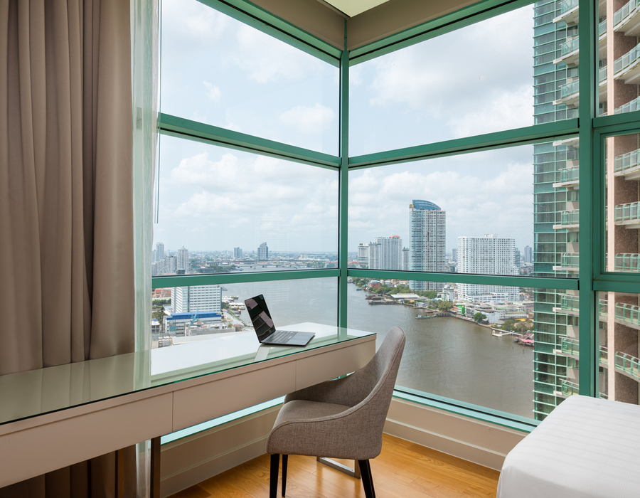Corner guestroom with floor-to-ceiling windows, river and city skyline views, glass-top work desk with laptop, modern chair and bed edge.