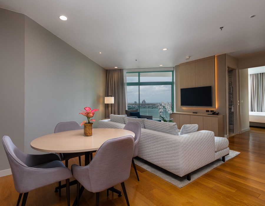 Modern suite with round dining table and floral centerpiece, plush sofa, wall TV, hardwood floors and waterfront city view
