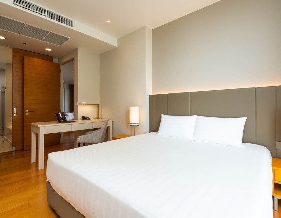 Modern hotel king room with large bed, padded headboard, bedside lamps, wood flooring, work desk with chair and open door to en-suite bathroom.