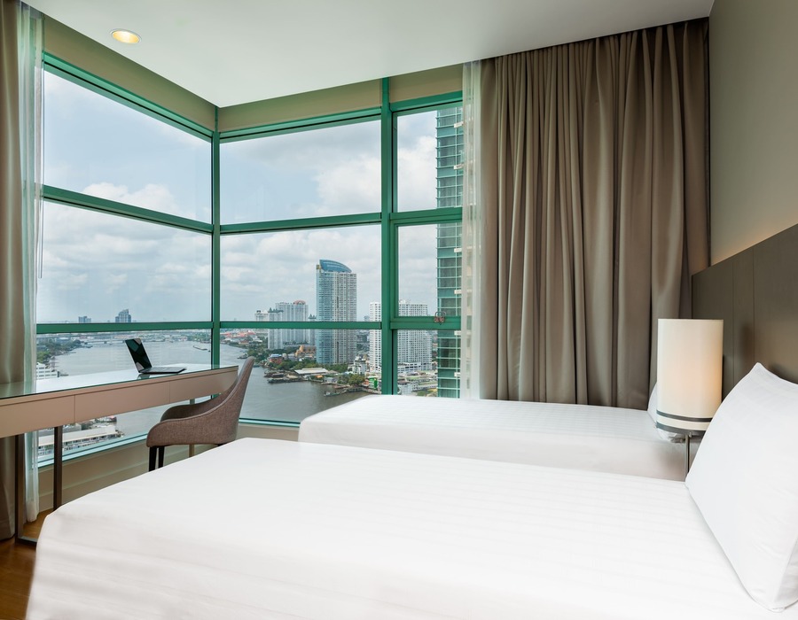 Contemporary twin room with floor-to-ceiling corner windows overlooking river and city skyline, work desk, soft neutral decor