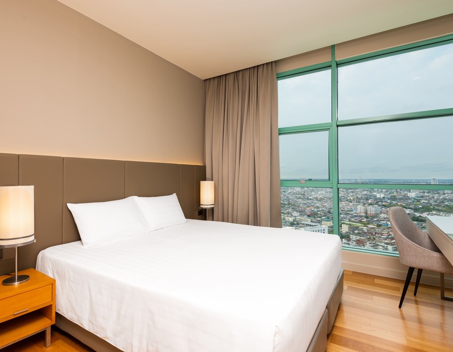 Modern hotel room with king bed, white linens, upholstered headboard, bedside lamps, desk and floor-to-ceiling city view window.