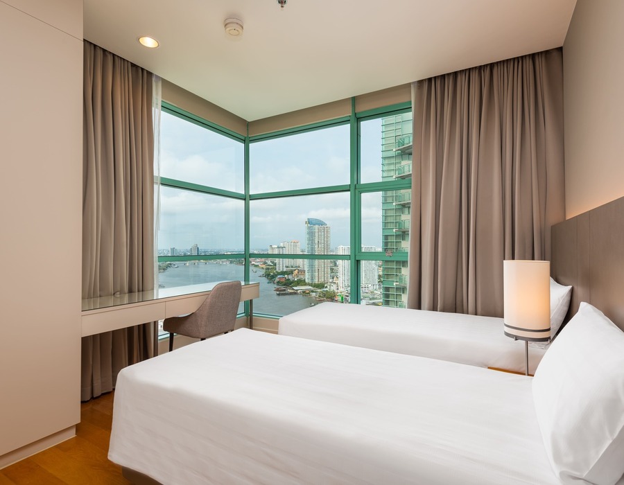 Twin room with two white beds, bedside lamp, desk by corner floor-to-ceiling windows overlooking river and city skyline.