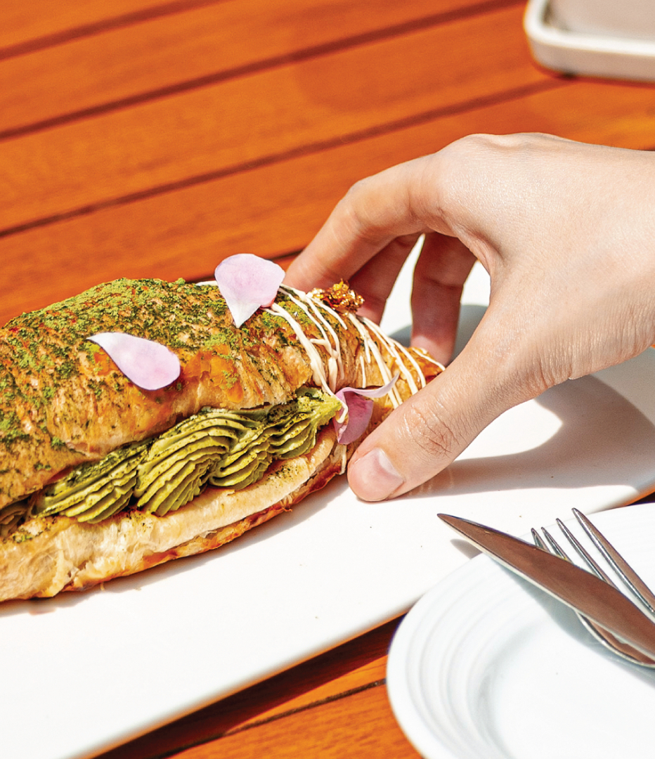 Hand reaching for a gourmet croissant filled with green cream and flower petals on a white plate at a wood table with cutlery