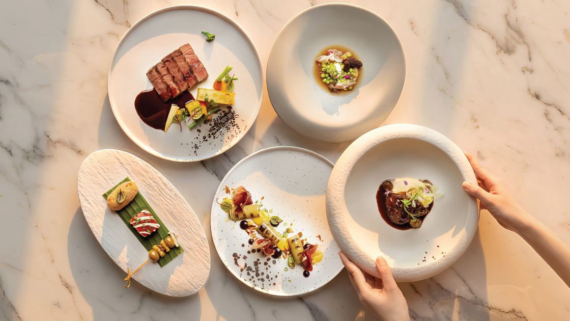 Fine-dining tasting menu: five artfully plated dishes on marble tabletop, steak and vegetable courses, hands placing bowl.