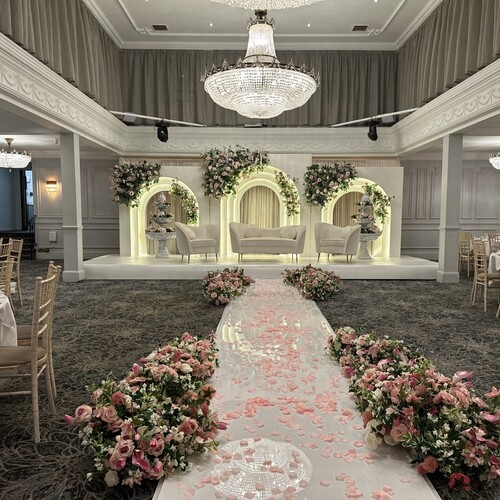 Elegant ballroom wedding with rose-petal aisle to illuminated arch stage, sofas, crystal chandeliers, pink floral displays.