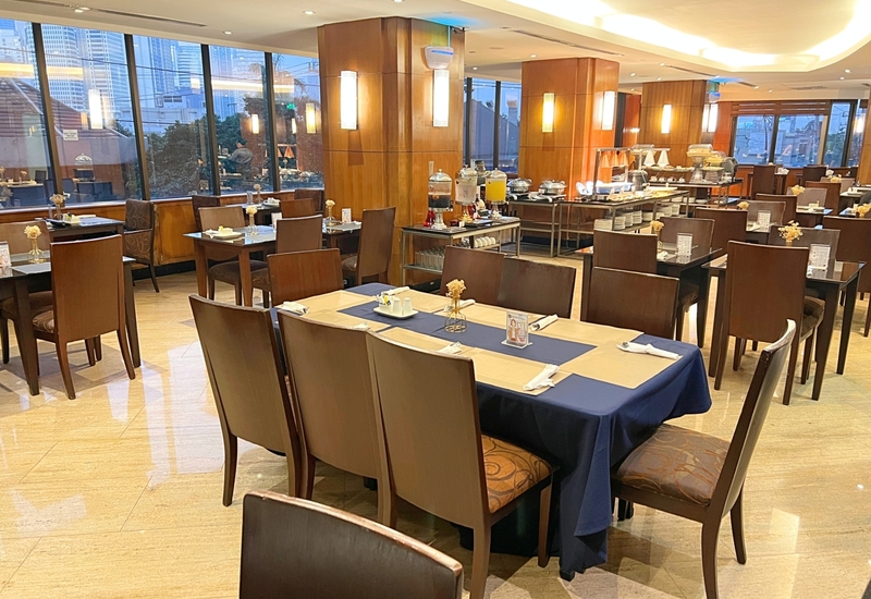 Bright hotel restaurant with wood-paneled columns, set tables with navy runners, self-serve breakfast buffet and city skyline through large windows.