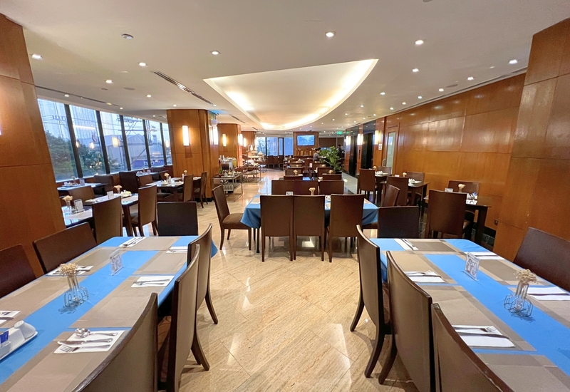 Spacious hotel dining room with blue table runners, wood-paneled walls, buffet station and floor-to-ceiling windows.