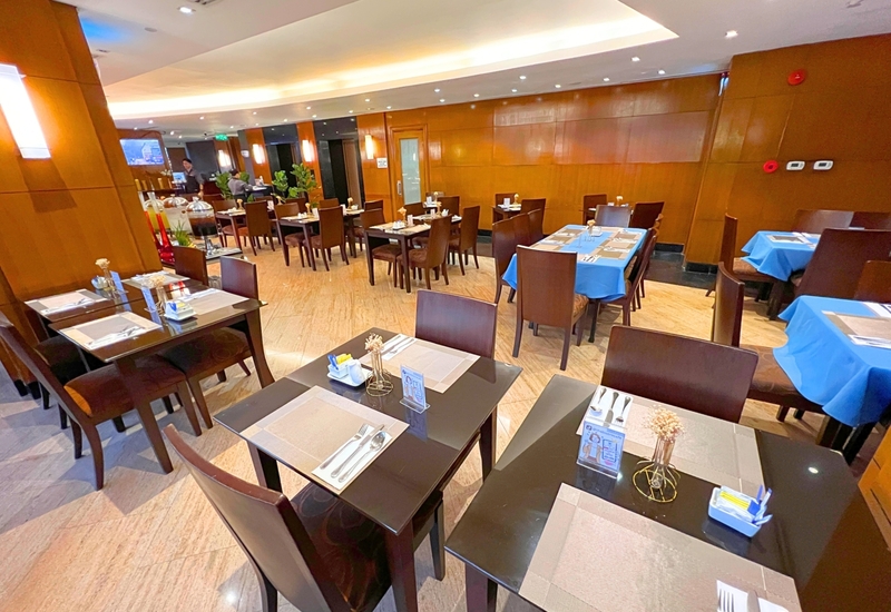 Hotel dining room with dark wood tables and chairs, set place settings, some tables with blue cloths and warm recessed lighting