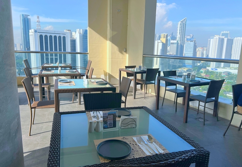 Rooftop hotel dining terrace with wicker tables and glass tops set for service, glass railing and panoramic city skyline view.