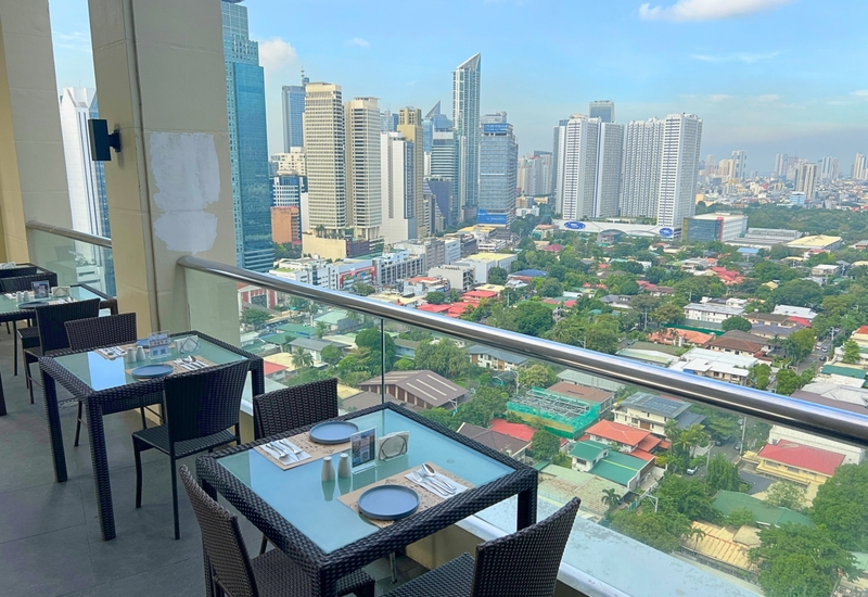 Hotel terrace dining with glass-top tables and wicker chairs overlooking downtown skyline and leafy neighborhood.