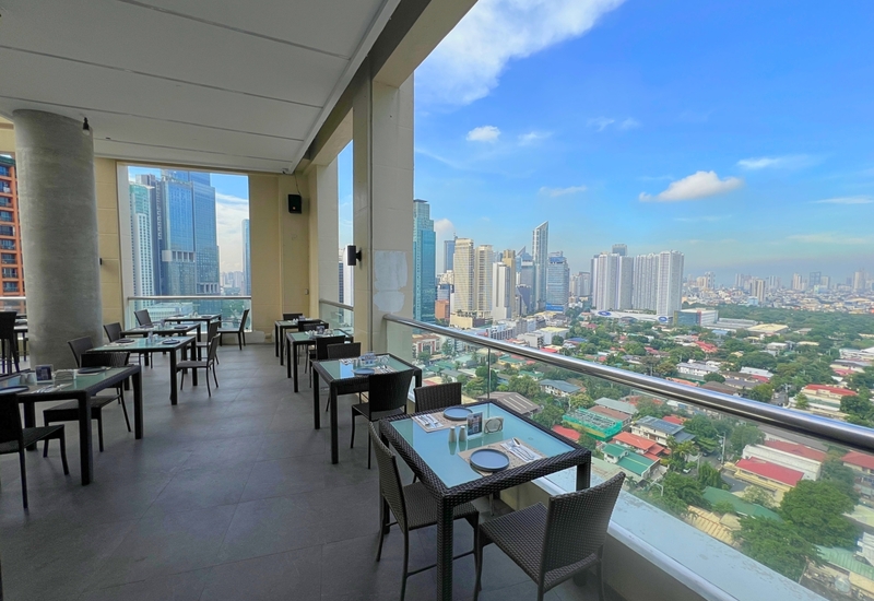High-floor open-air hotel terrace restaurant with glass railing, set tables and wicker chairs overlooking panoramic city skyline.