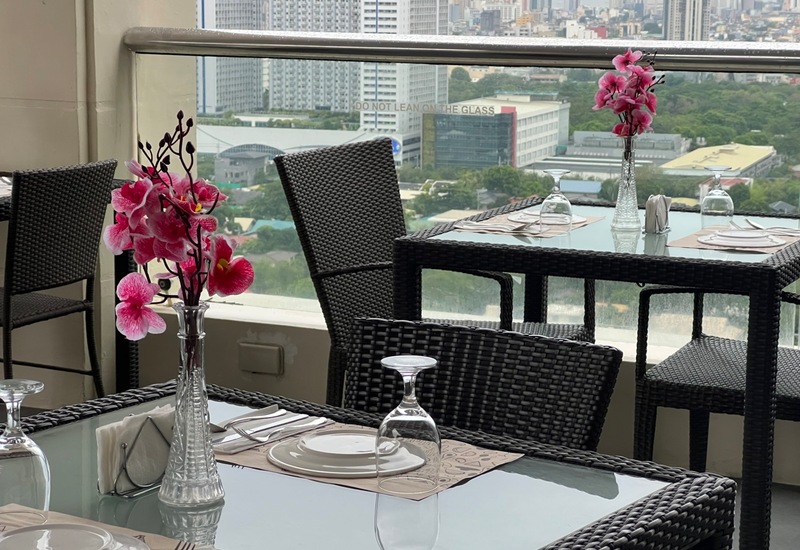 Rooftop hotel dining with wicker tables, glass tops, pink orchid centerpieces and set plates, overlooking city skyline.