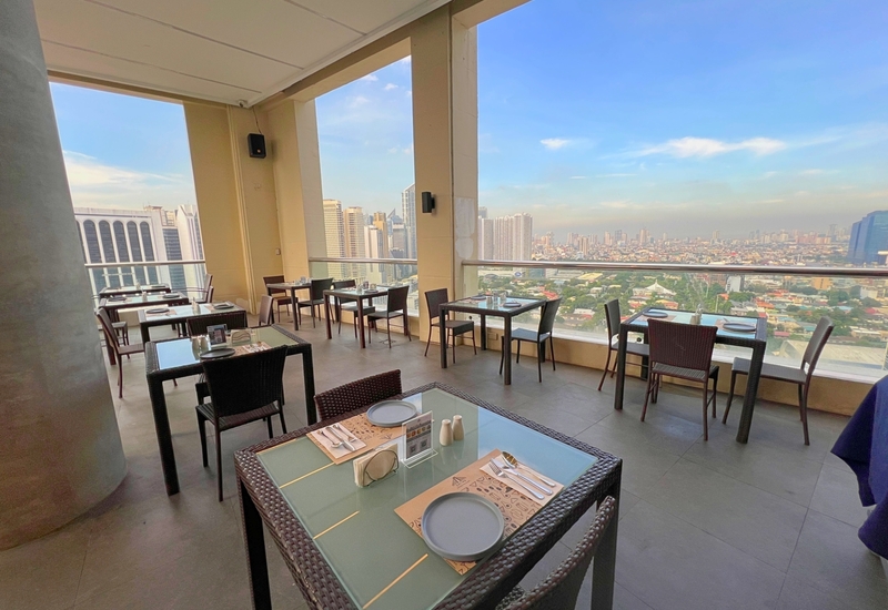 Rooftop hotel dining terrace with set tables, wicker chairs and glass railing offering panoramic city skyline views.
