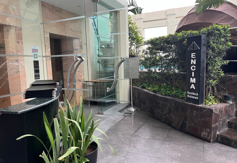Glass entrance to Encima roofdeck restaurant with curved metal handles, stacked menus on granite desk, plants and pool glimpse.