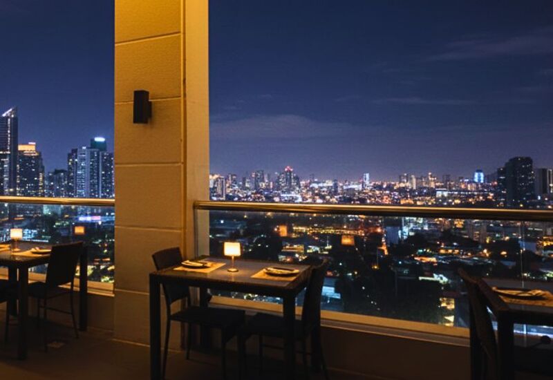 Rooftop hotel restaurant terrace with candlelit tables, glass railing and sweeping city skyline views at night.