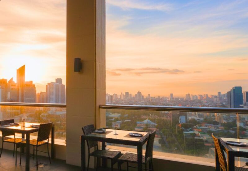 Rooftop hotel terrace restaurant with set dining tables, glass railing and panoramic city skyline at golden sunset.