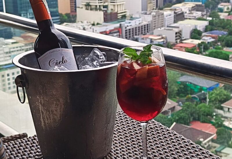 Hotel balcony table with chilled red wine in ice bucket and fruit-filled sangria glass garnished with mint, city skyline view.