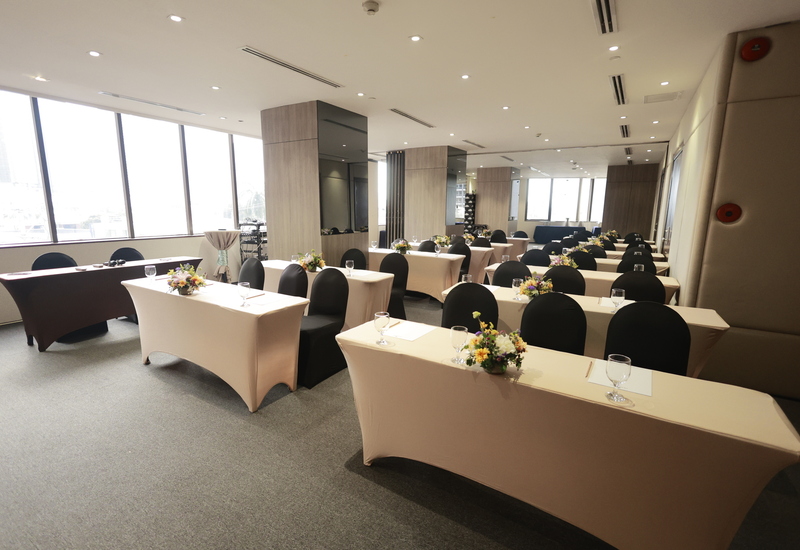 Bright meeting room with floor-to-ceiling windows, beige draped classroom-style tables, black chairs, floral centerpieces and water glasses.