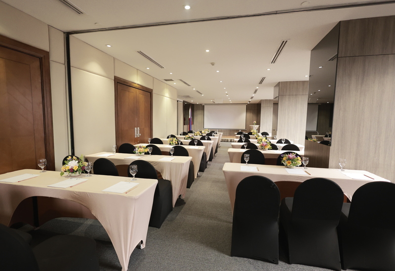 Conference room set classroom-style with beige-covered tables, black chair covers, floral centerpieces, water glasses and front projection screen.
