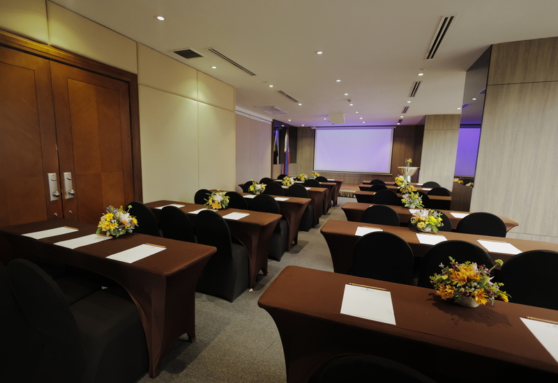 Hotel conference room set classroom-style with brown tables, black chairs, floral centerpieces and projector screen.