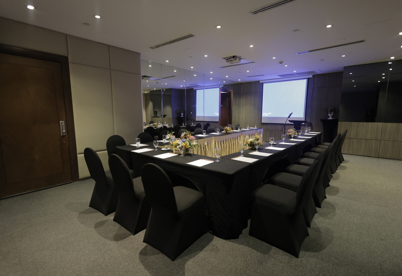 Hotel boardroom with U-shaped black-draped tables, chairs, floral arrangements, notepads, water glasses and projector screen.