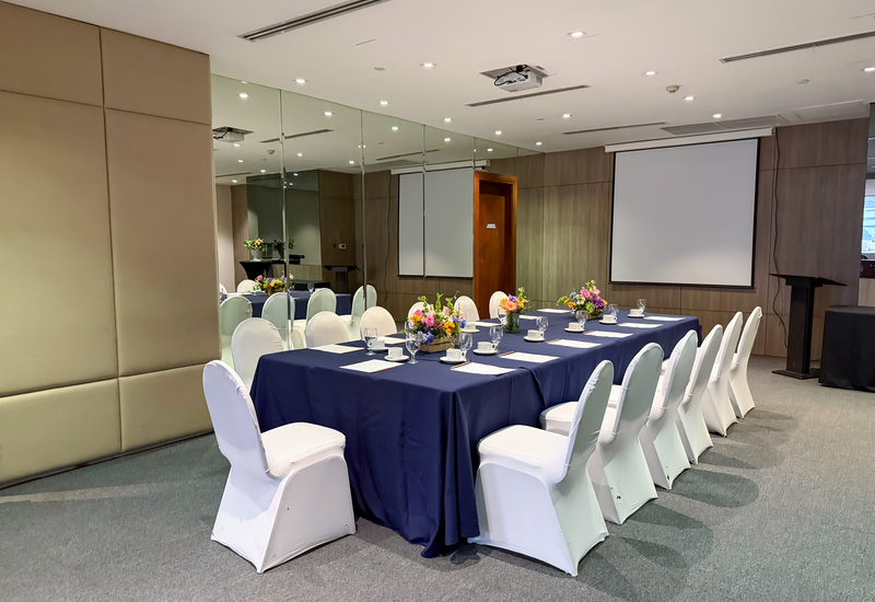 Boardroom-style meeting room with U-shaped navy-draped table, white chair covers, floral centerpieces, projector screen and podium for presentations