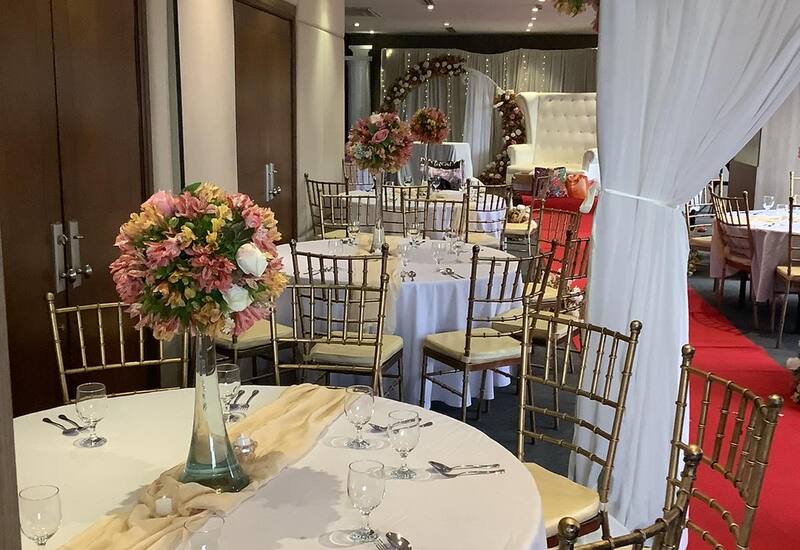 Elegant wedding reception room with round tables, white linens, gold Chiavari chairs, floral centerpieces, red aisle carpet and tufted white loveseat.