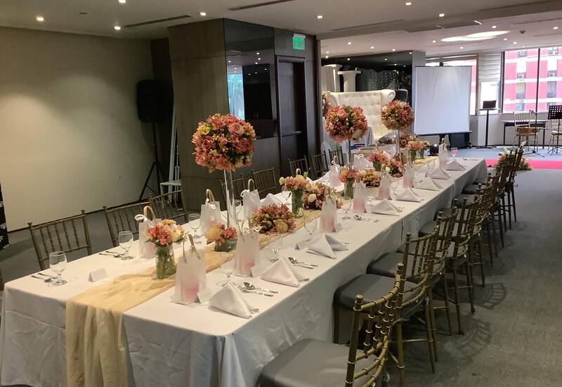 Long banquet table set for a wedding reception with white linens, gold chiavari chairs, pink floral centerpieces, gift bags.