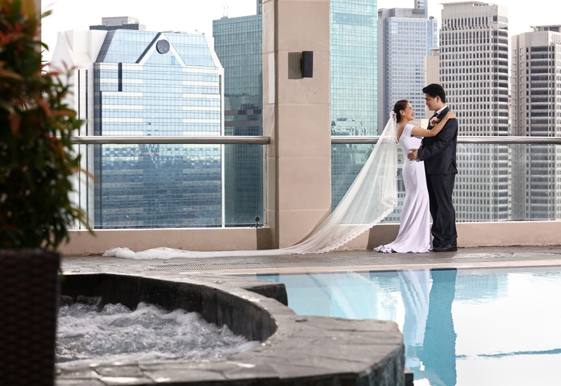 Bridal couple embrace on a hotel rooftop terrace beside pool and jacuzzi, romantic city skyline view.