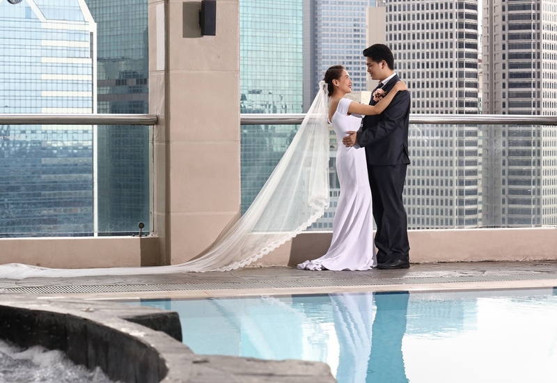 Newlyweds embracing on a rooftop hotel terrace by a pool, bride’s long veil trailing with city skyscraper skyline view