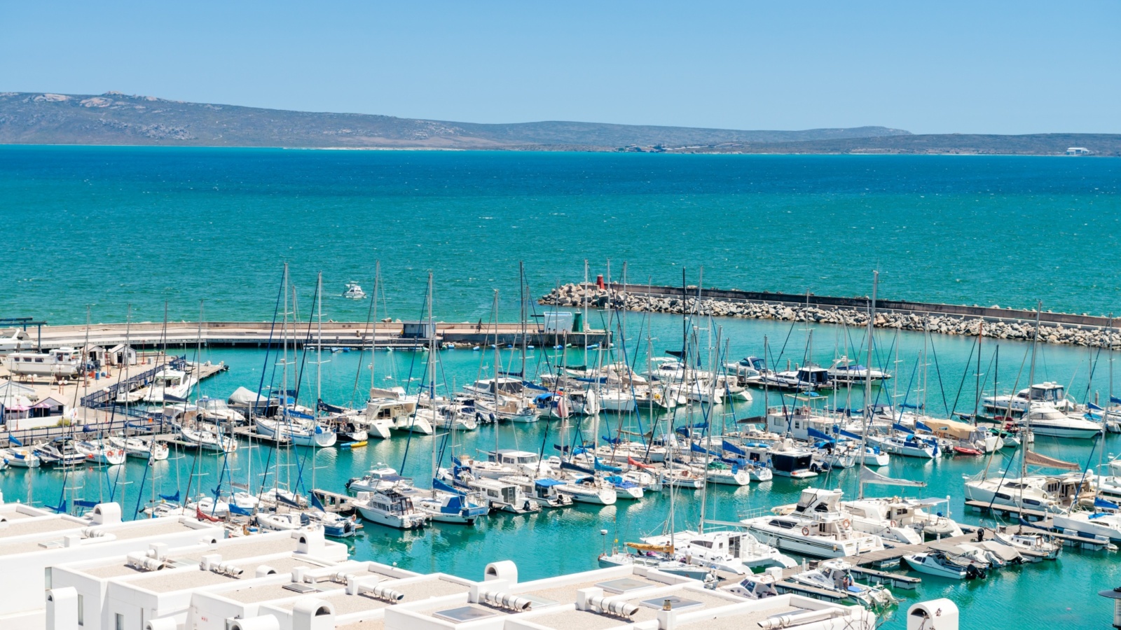Marina of moored yachts and sailboats, turquoise sea and distant headland, white hotel terraces in foreground.