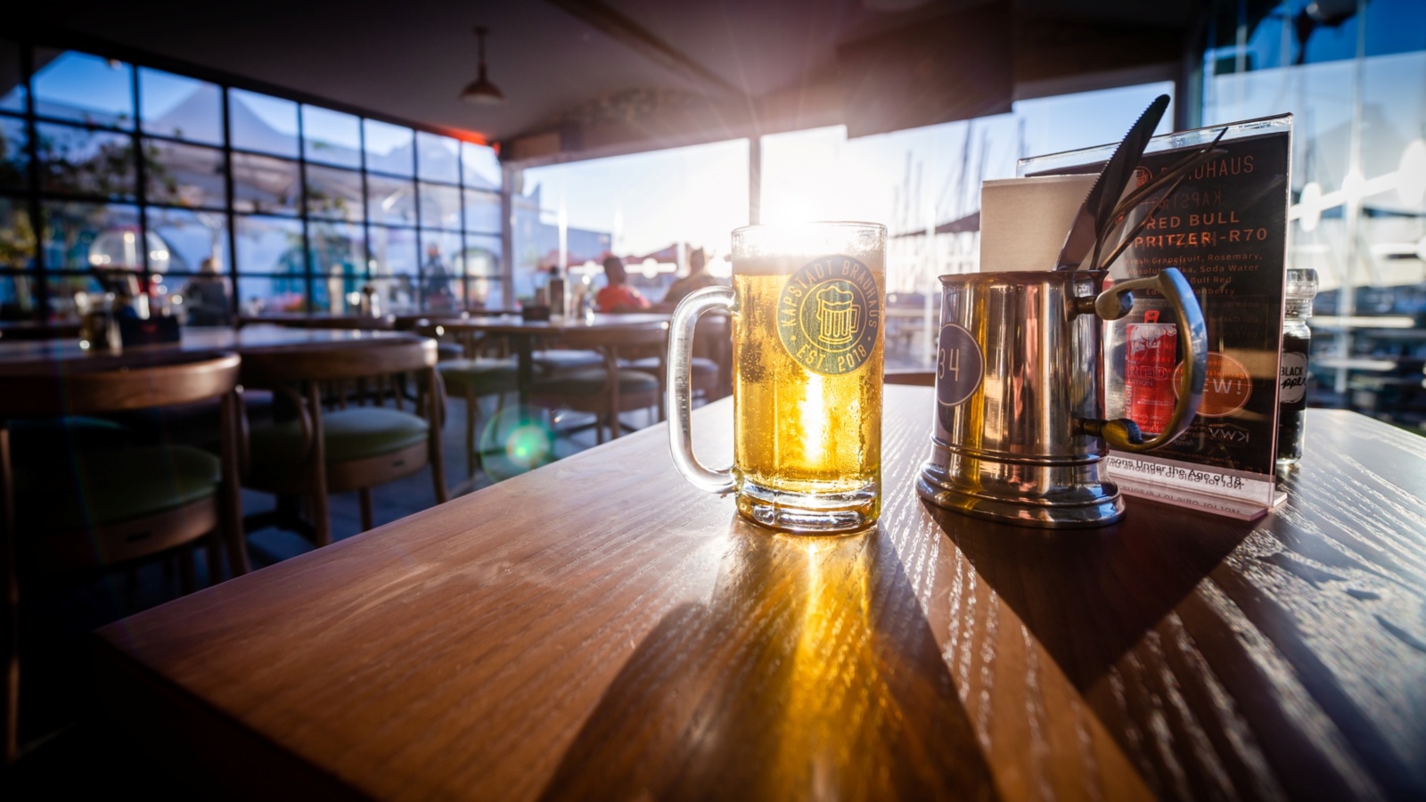 Sunlit beer mug on a wooden table in a waterfront restaurant, metal cutlery holder and menu, warm relaxed atmosphere.