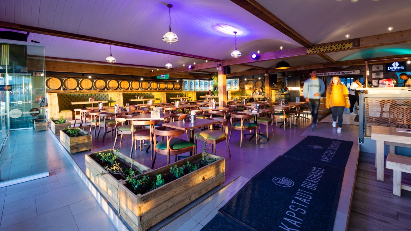 Brewery-style dining area with barrel-lined wall, wooden tables, planter boxes, purple lighting and guests at the bar.