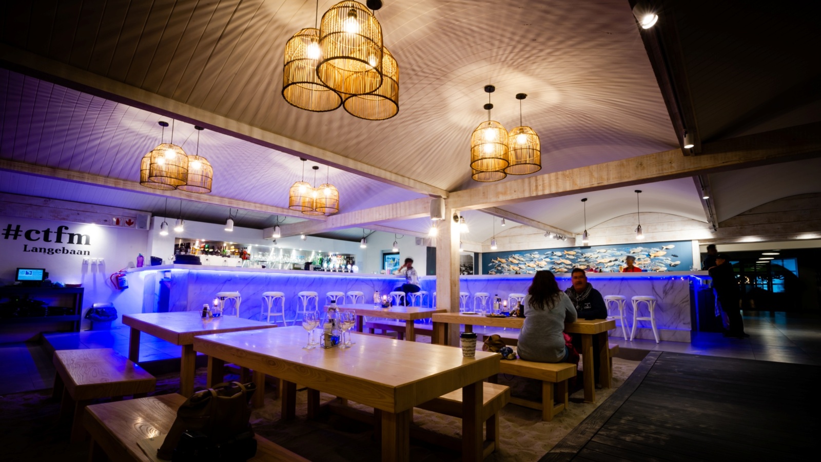 Casual beachfront bar and restaurant with communal wooden tables and benches, blue-lit marble bar, rattan pendant lights, couple dining, fish wall art.