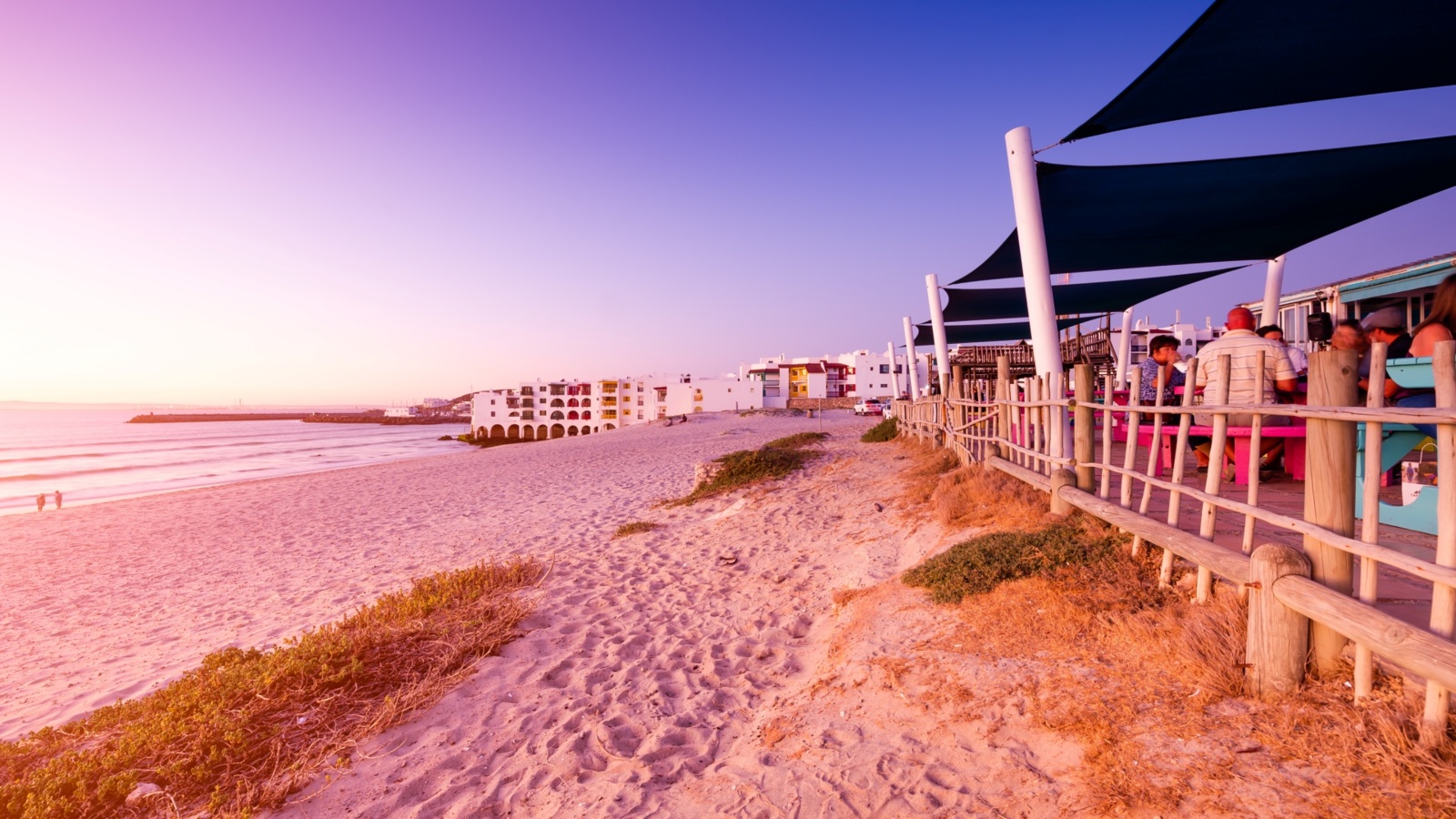 Beachfront restaurant patio with shaded seating and diners beside a sandy path leading to white arch-fronted seaside hotel at sunset.