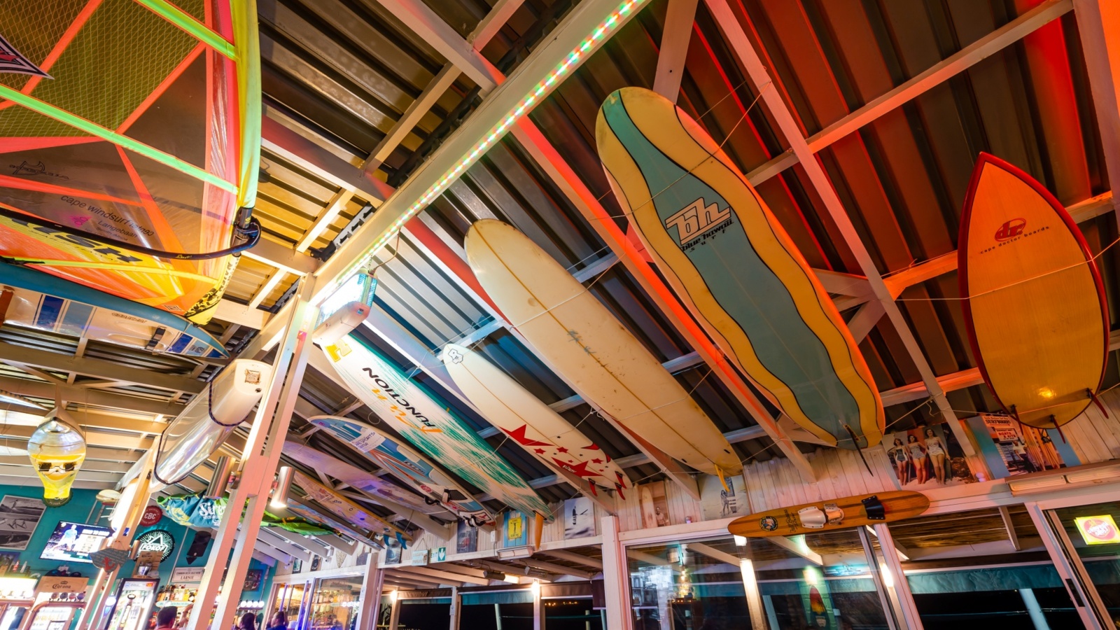 Beachfront bar interior with surfboards and sails suspended from exposed timber beams, colorful LEDs, lively dining.