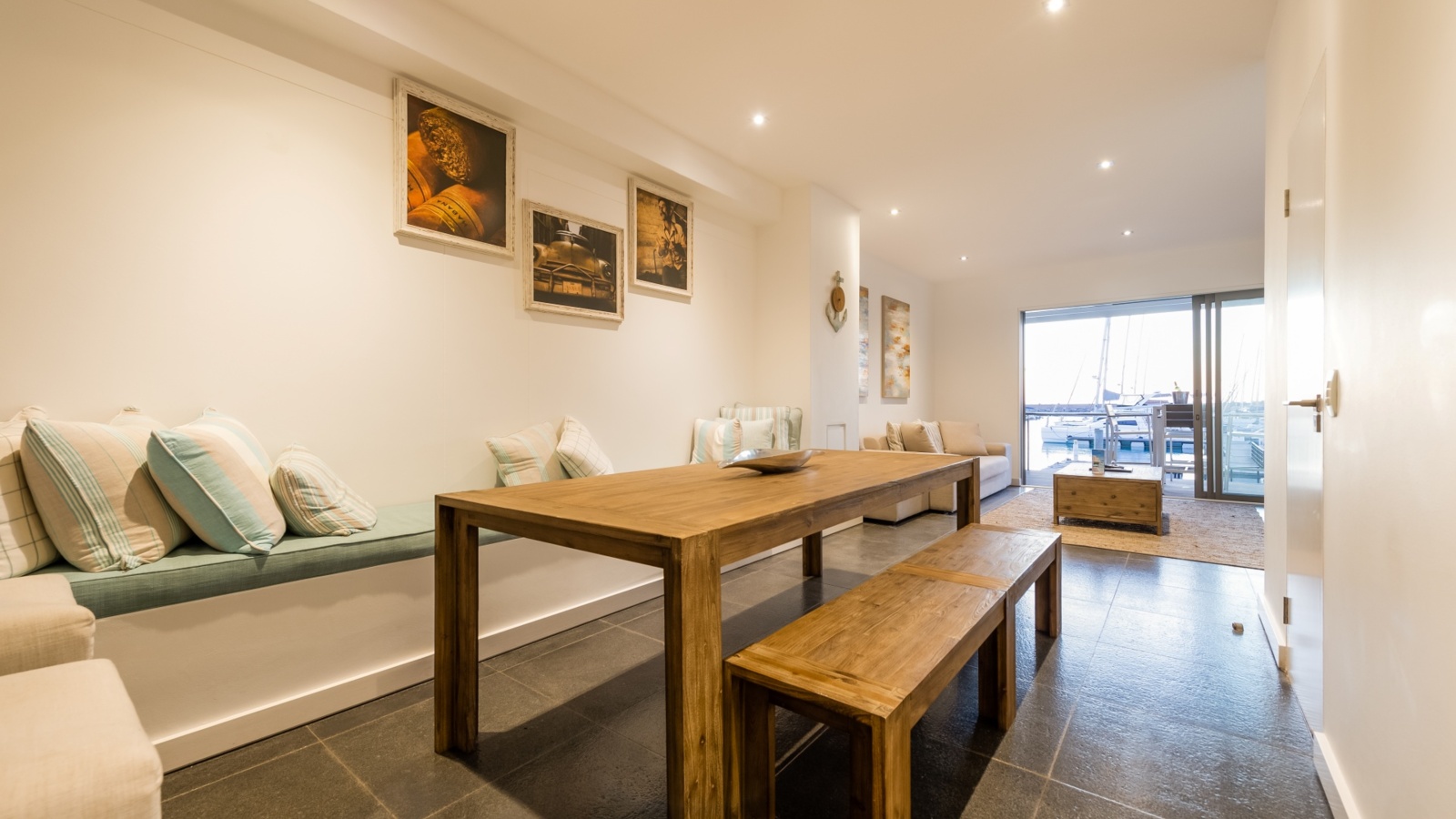 Coastal-style dining and lounge with long wooden table and benches, cushioned banquette and sofa, sliding doors to marina view.