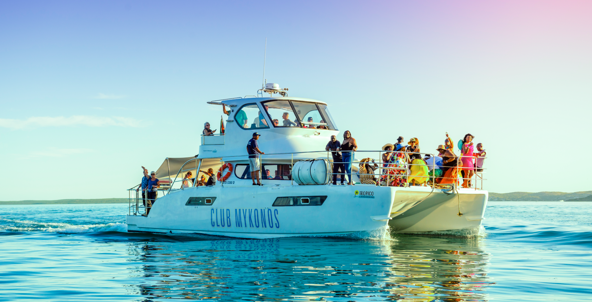 Daytime party catamaran cruise with lively guests on upper deck, drinks and dancing, calm turquoise sea and distant shoreline.