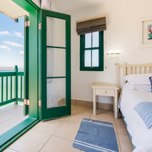 Seaside room with green-framed French doors opening to a balcony over turquoise water, white bedding and striped cushions.