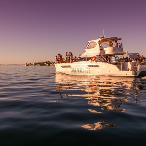 Sunset cruise yacht with guests on deck, coastal villas along shore, calm reflective water and warm twilight sky
