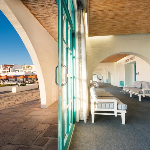 Shaded hotel colonnade with turquoise doors and striped lounge benches facing waterfront dining with orange umbrellas.