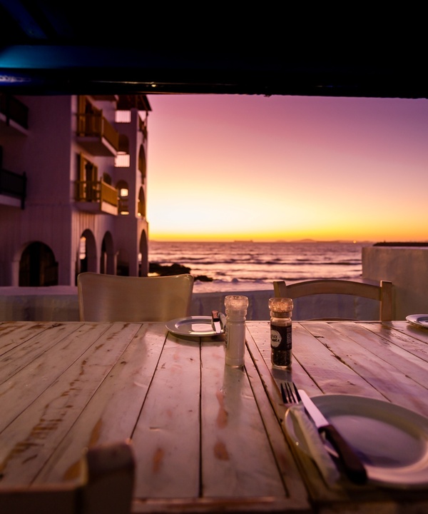 Covered beachfront terrace with rustic wooden table set for four, salt-and-pepper shakers, arched hotel balconies and a vivid sunset over the ocean