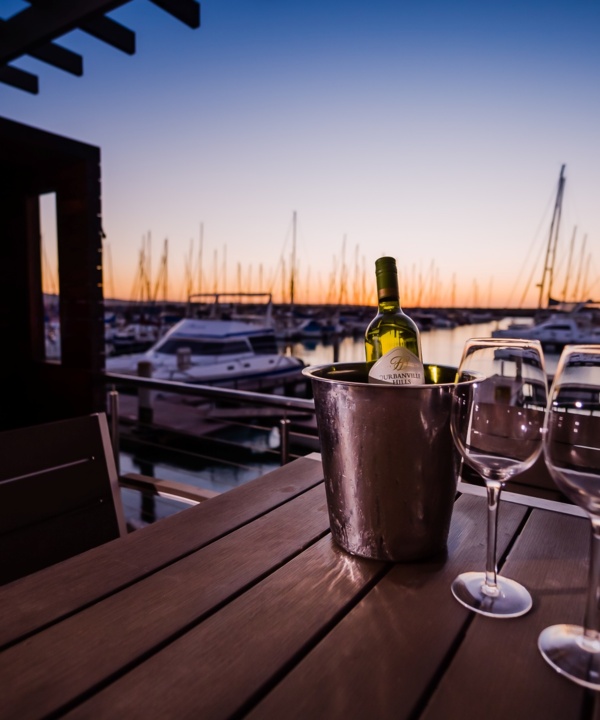 Waterfront terrace table with chilled bottle in an ice bucket and two wine glasses, overlooking yachts at sunset in a marina.