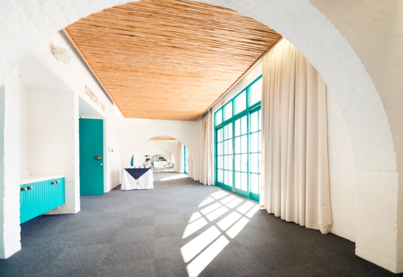 Sunlit hotel event corridor with white arches, bamboo ceiling, turquoise French doors, carpeted floor and refreshment table.