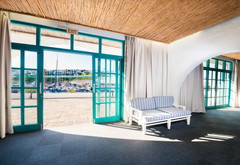 Coastal hotel lounge with turquoise doors opening to a marina, striped daybed, bamboo ceiling, arched white walls